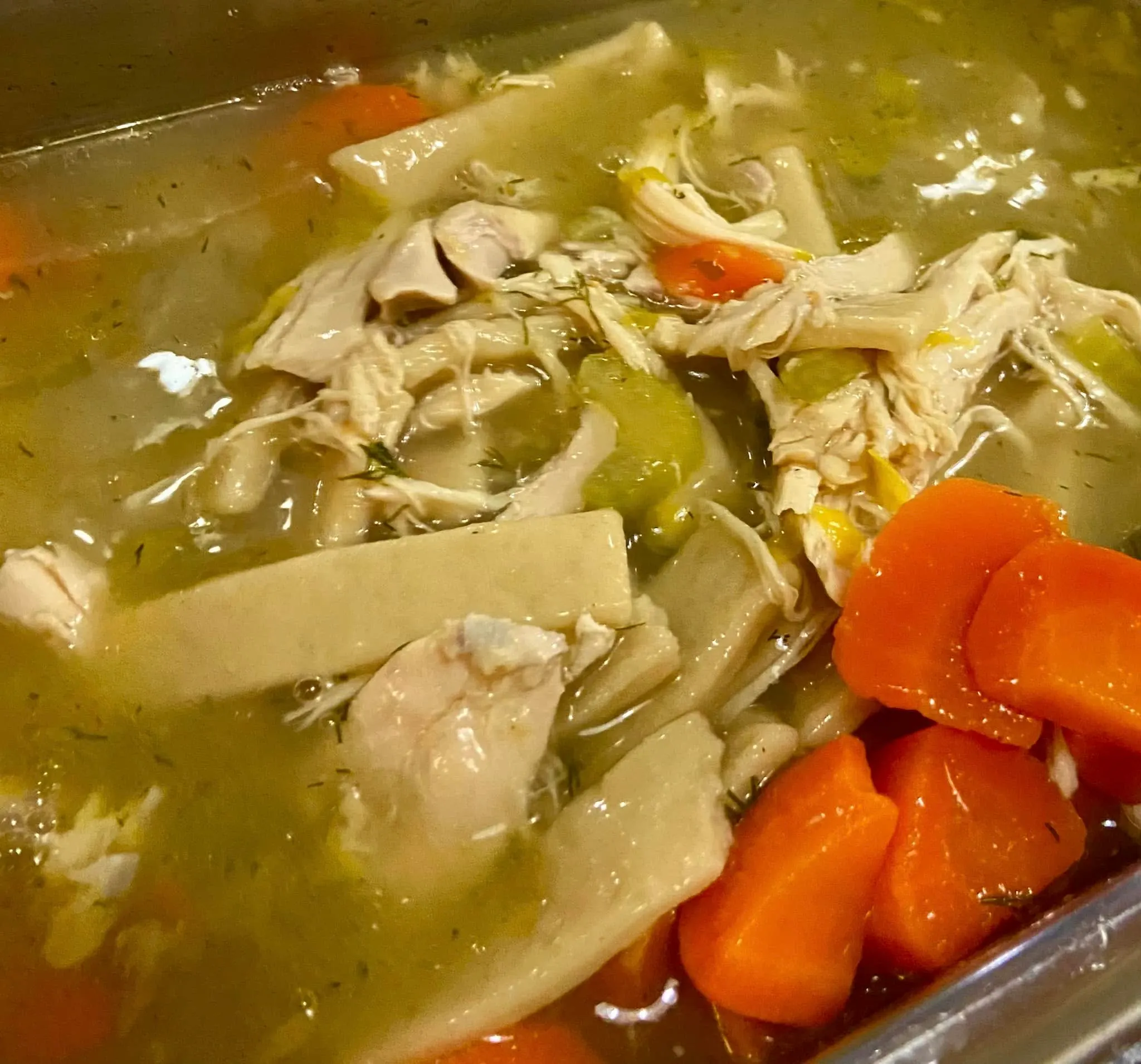 A pot of chicken noodle soup with wide egg noodles, shredded chicken and carrots